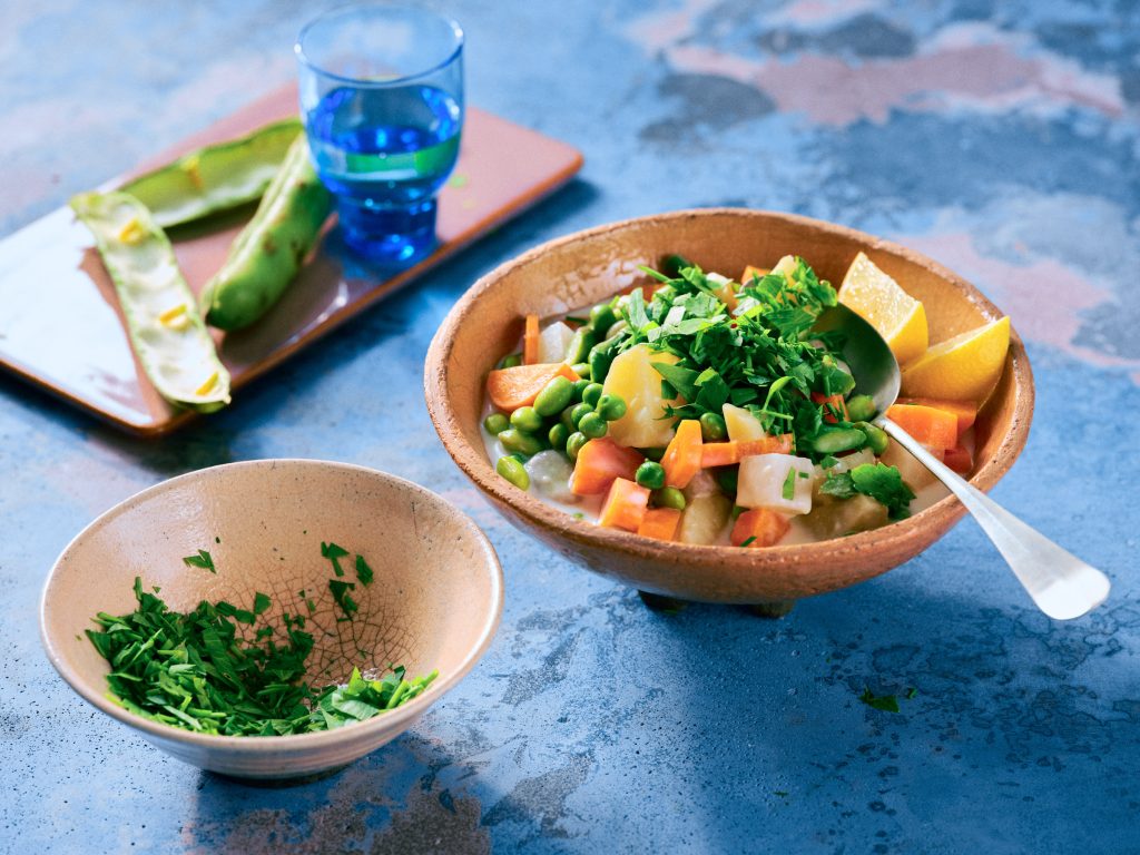 Totale von TASTE APPEAL– Rezept: Schnüsch mit Edamame in Terracottaschüssel auf blauem Untergrund, dahinter leere Edamameschoten, im Vordergrund kleine Schüssel mit Kräutern