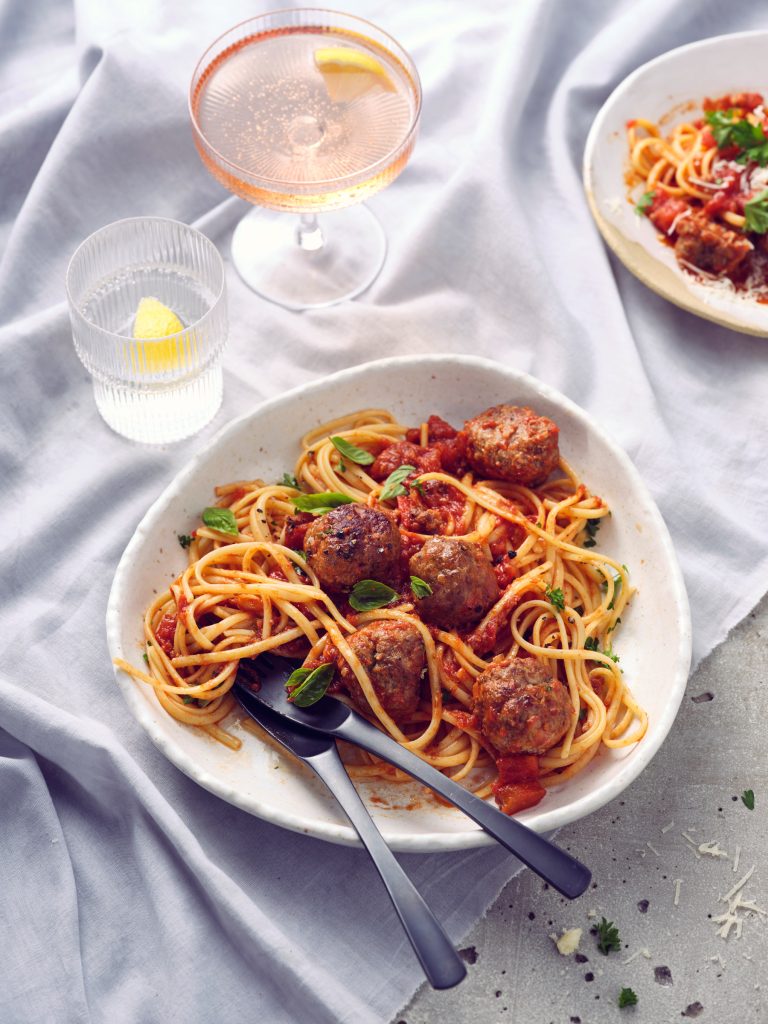 Totale von TASTE APPEAL– Rezept Grandmas Meatballs mit schwarzem Besteck auf heller Tischdecke, daneben Gläser mit Drink