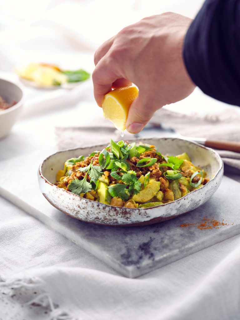 Totale von TASTE APPEAL– Rezept Schmorgurkencurry mit Sonnenblumenhack in Schüssel, daneben Besteck, im Hintergrund kleiner Teller mit Zitrone, Currypuler und Frühlingszwieblen, Hand drückt über Gericht Zitrone aus