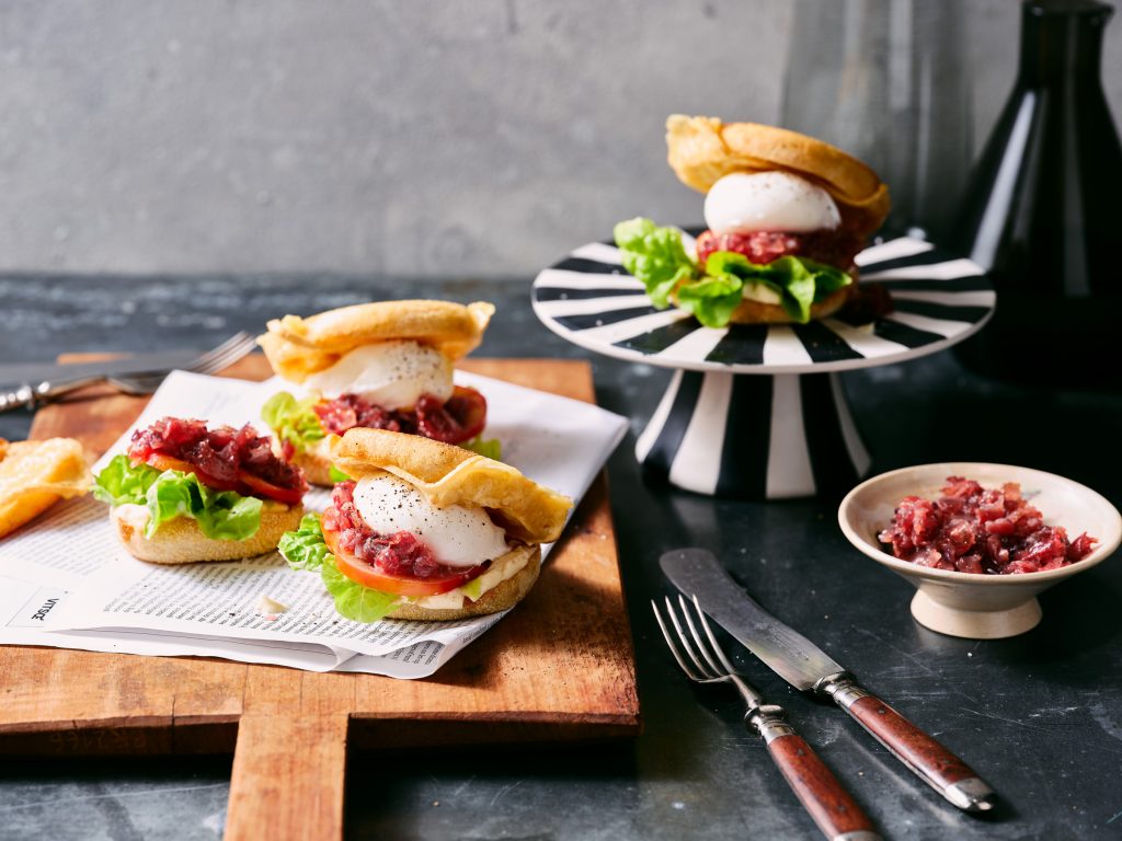 Totale von TASTE APPEAL– Rezept Breakfast-Burger mit Deichkäse und pochiertem Ei auf gestreiften Podest, neben auf Zeitungspapier und Holzer drei weitere, davor Besteck und Bavonmarmelade in Schüssel