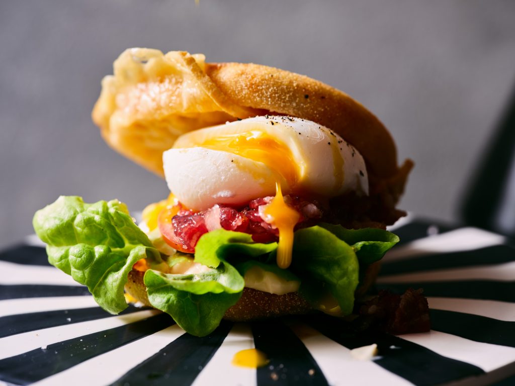 Großaufnahme von TASTE APPEAL– Rezept Breakfast-Burger mit Deichkäse und pochiertem Ei auf gestreiften Podest