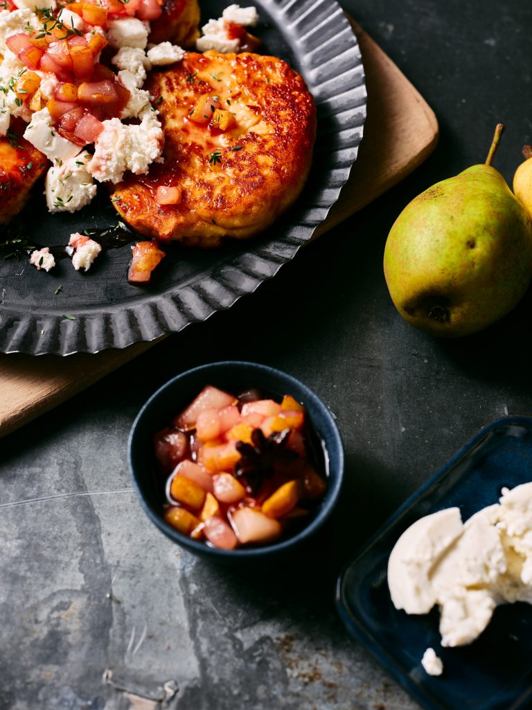 Nahaufnahme von TASTE APPEAL– Rezept Kartoffel Küchlein mit Ziegenfrischkäse und Birne auf schwarz Teller, daneben in kleiner Schüssel Topping und Birne