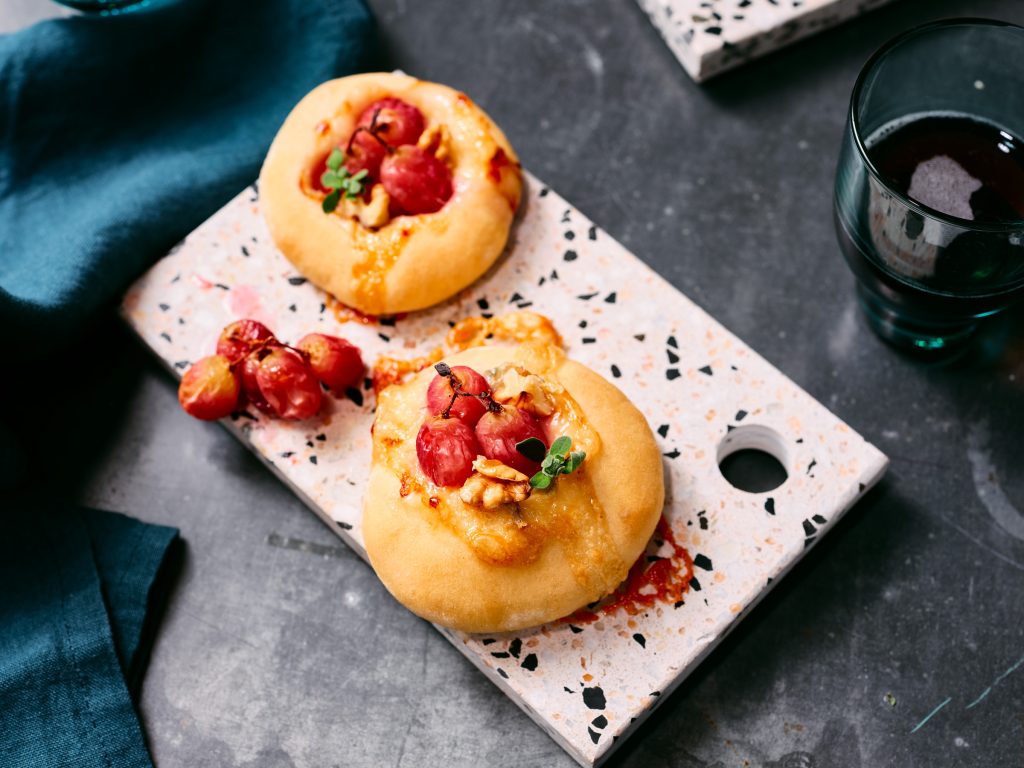 Nahaufnahme von TASTE APPEAL– Rezept Mini-Pizzen mit Gorgonzola, Trauben und Walnüssen auf Steinplatte, daneben Gläser und blaue Tücher