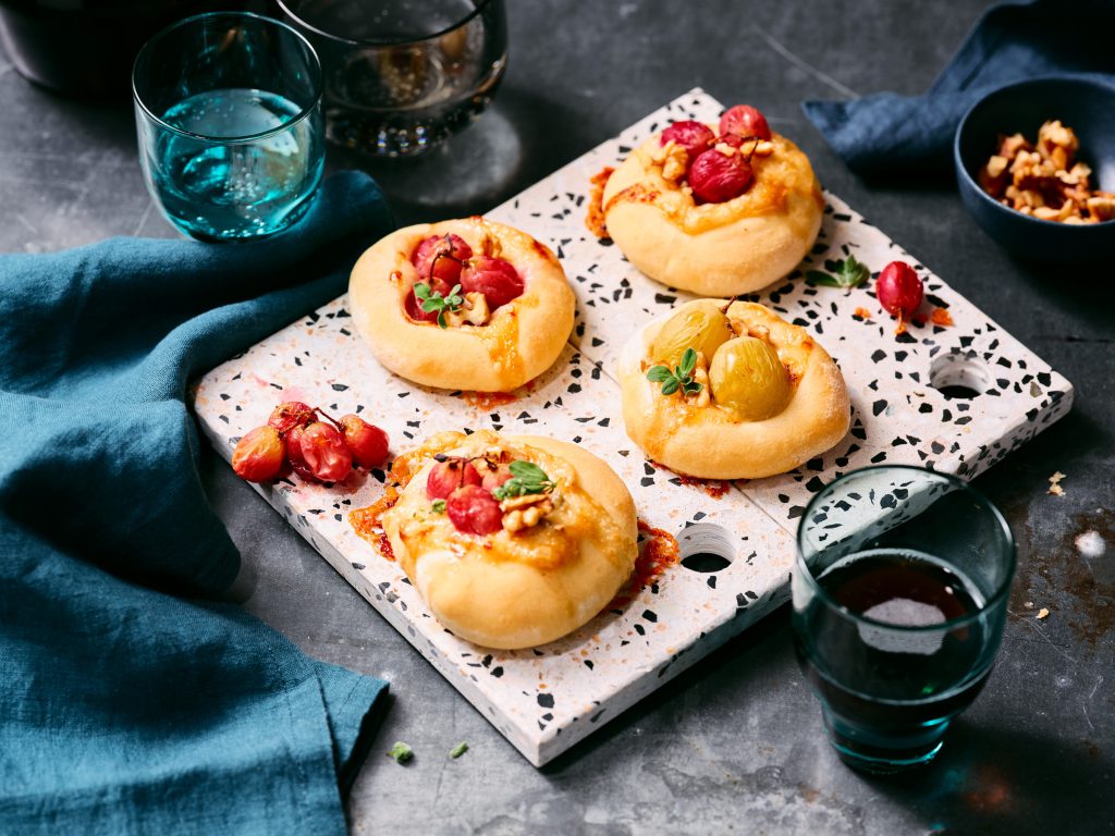 Totale von TASTE APPEAL– Rezept Mini-Pizzen mit Gorgonzola, Trauben und Walnüssen auf Steinplatte, daneben Gläser und blaue Tücher