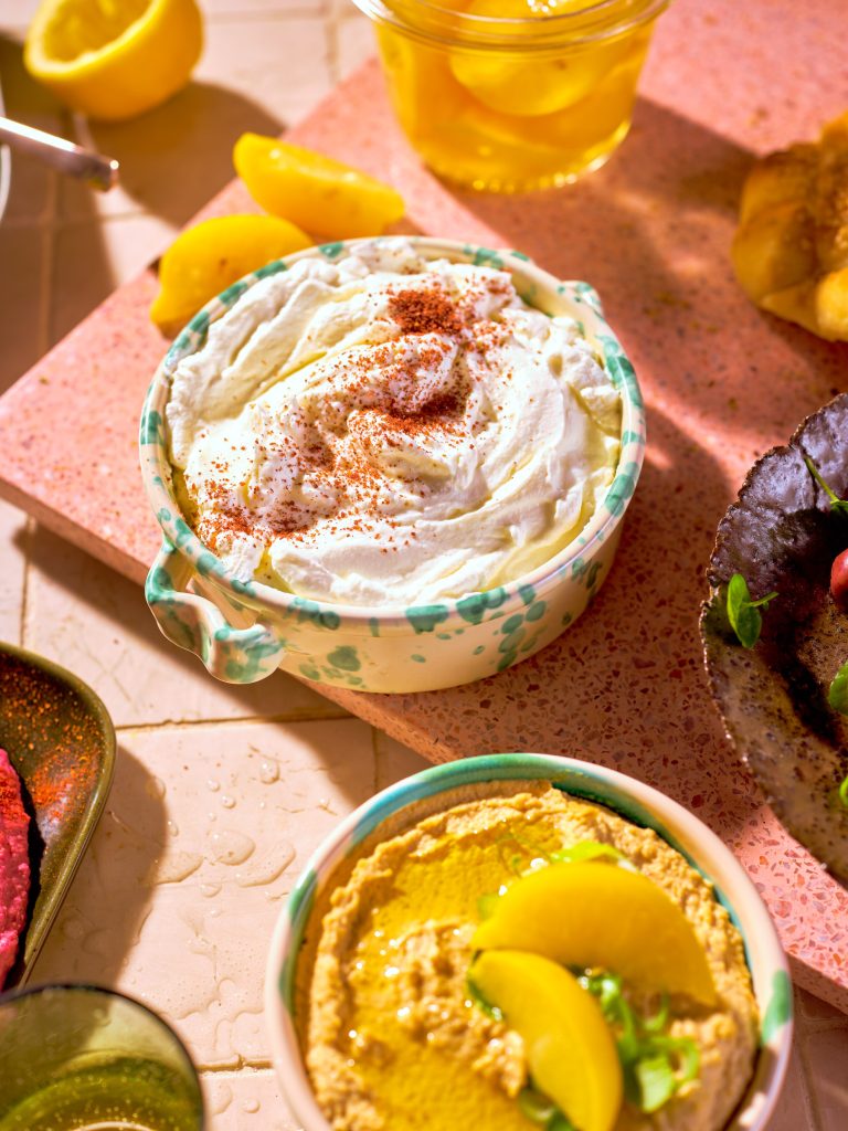 Totale von TASTE APPEAL– Rezept Labneh Frischkäse-Joghurt auf pink Steinplatte, neben Oliven in Schüssel, Hummus, Zitronen