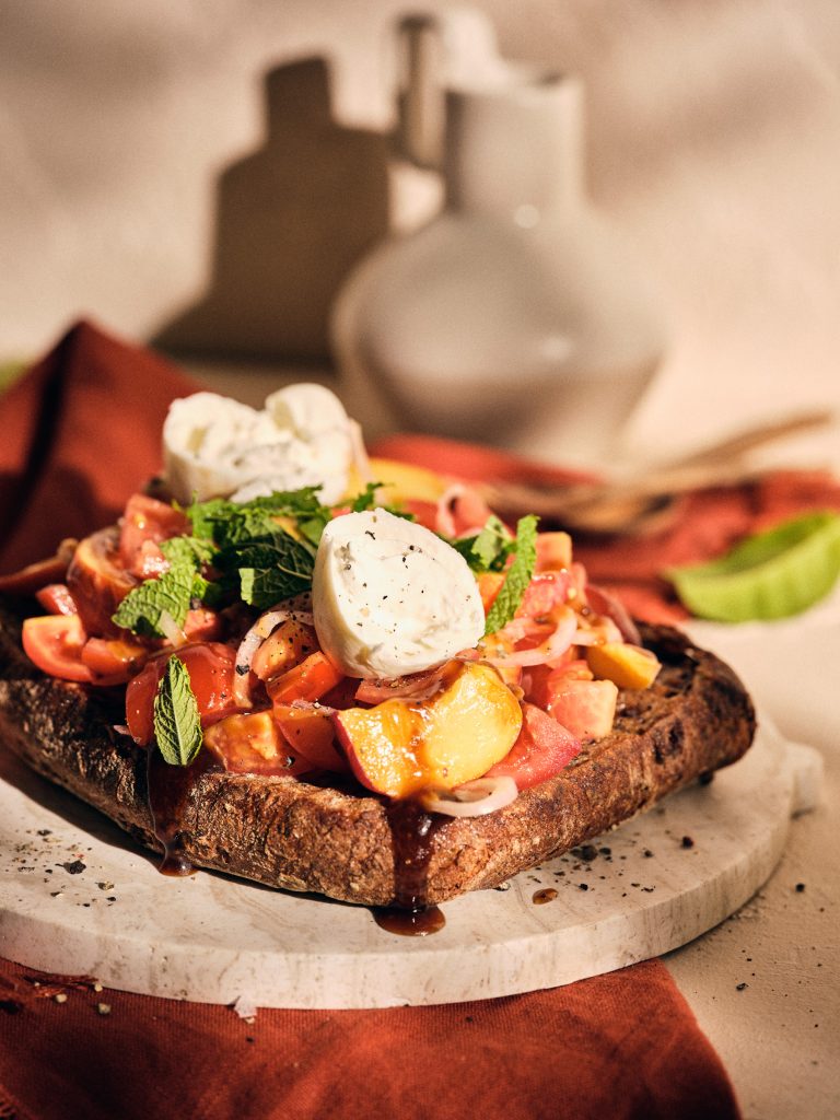 Halbtotale von TASTE APPEAL– Rezept Tomaten-Brotsalat mit Pfirsich und Burrata auf Steinboden mit Tuch