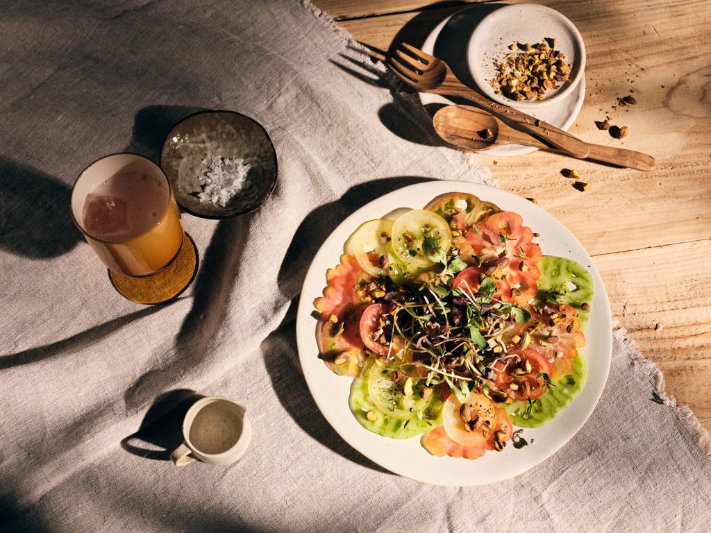 Totale von TASTE APPEAL– Rezept Tomaten-Carpaccio mit Basilikum-Öl und gerösteten Pistazien auf weißem Teller, im Hintergrund kleiner Teller mit Besteck und Pistazien, daneben Getränk und Salz