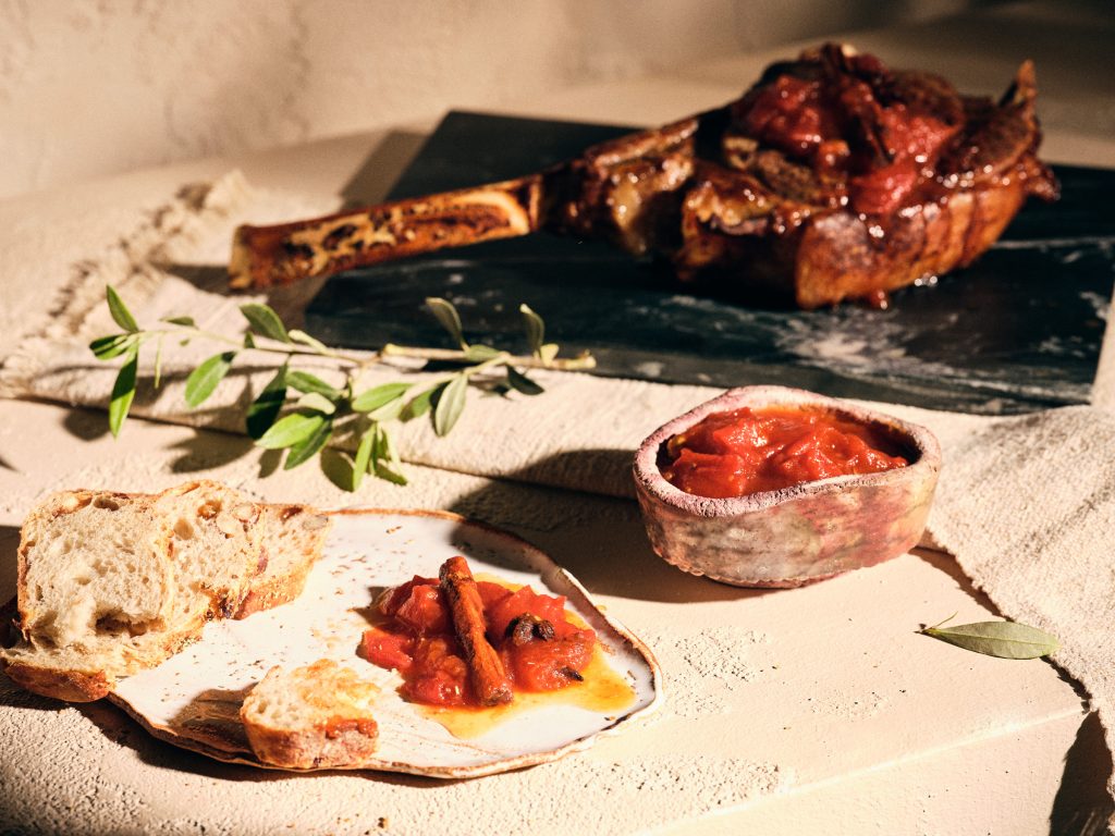 Halbtotale von TASTE APPEAL– Rezept Tomaten-Chutney mit Vanille, Sternanis und Chili auf Keramikteller, daneben Brotscheibe, im Hintergrund Fleischstück auf Steinplatte