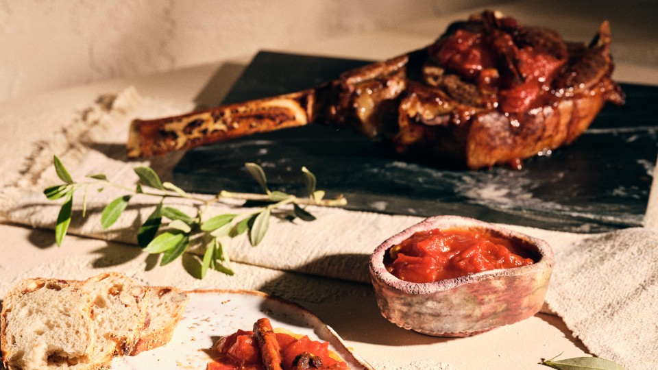 Halbtotale von TASTE APPEAL– Rezept Tomaten-Chutney mit Vanille, Sternanis und Chili auf Keramikteller, daneben Brotscheibe, im Hintergrund Fleischstück auf Steinplatte
