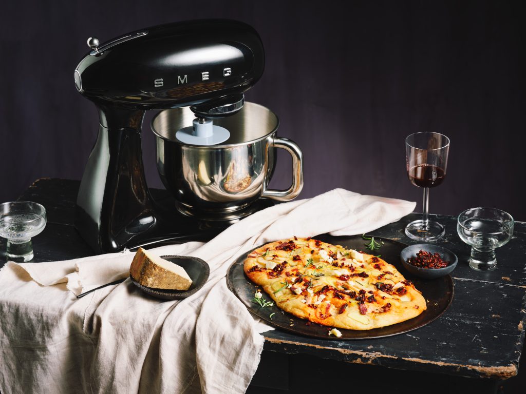 TASTE APPEAL- Rezept, Kartoffel Focaccia mit Chili auf schwarzem Teller, daneben sieg Küchenmaschine, Weinglas, Käse und helle Tischdecke