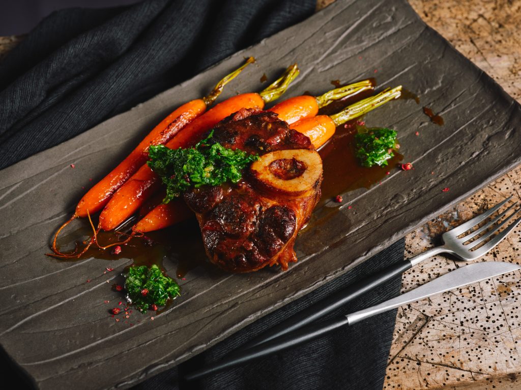 Nahaufnahme TASTE APPEAL- Rezept, Ossobuco mit Karamellisierten Karotten und Gremolata auf grauer Platte, daneben Besteck