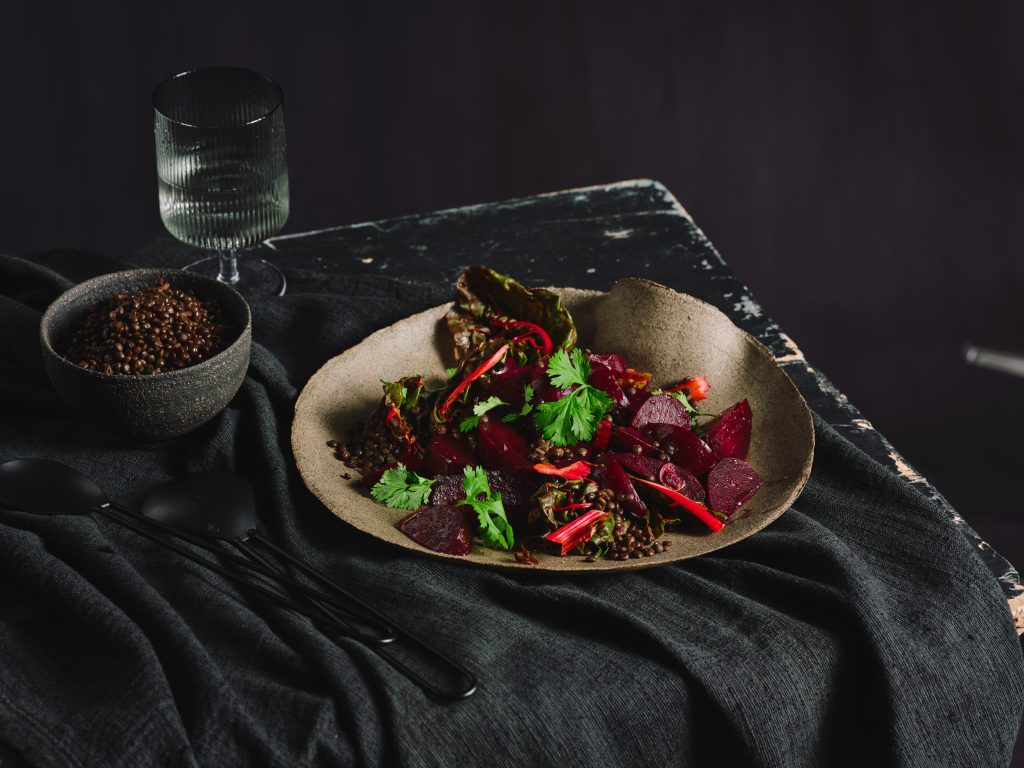 TASTE APPEAL- Rezept, Rote-Bete-Salat mit Ingwer und Koriander auf geschwungenem Teller auf dunkler Tischdecke, im Hintergrund Glas und kleine Schale